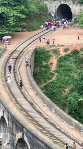 The video opens with a wide shot of a curved railway track leading towards a tunnel entrance. Lush green trees and vegetation surround the area, with a steep, earthy embankment to the right of the tracks. Several people are scattered along the railway embankment and near the tunnel entrance. On the left side of the tracks, a stone viaduct is partially visible at the bottom of the frame.

As the video progresses, the camera angle remains relatively static, offering a consistent view of the scene. Several individuals can be seen walking along the railway tracks and the paths adjacent to them. Near the tunnel, a group of people have gathered, some standing and others appearing to be walking towards or away from the tunnel entrance. A person in a yellow shirt is visible on the right side of the tunnel, near a sign.

A small stall with a red and white umbrella is set up to the left of the tracks, near the beginning of the curve. Further down the tracks, individuals are walking in both directions. One person in a white dress and another in a dark outfit are seen walking on the viaduct-like structure beside the tracks.

The overall impression is a scenic railway line in a natural setting, with tourists or locals present at various points, particularly near the tunnel, suggesting it might be a point of interest or a transit area. The lighting suggests it is daytime, with overcast conditions contributing to soft shadows.