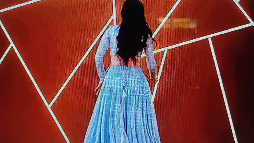 A woman with dark hair, wearing a light blue, sparkling crop top and a matching long skirt, dances in front of a textured brown wall with a geometric pattern of white lines. She holds the sides of her skirt and moves her arms in a fluid motion, her mouth open as if singing or exclaiming. The lighting is bright, highlighting the sparkle of her outfit.