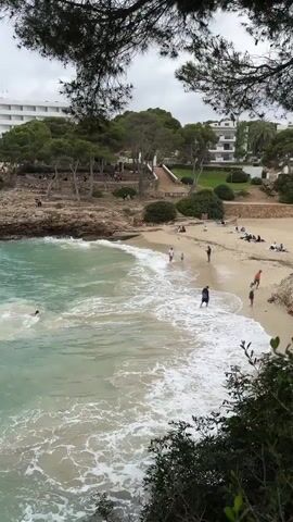 The video presents a high-angle shot of a beach on what appears to be a cloudy day. The water is a clear, light turquoise color near the shore, deepening to a darker hue further out. Waves are visibly crashing onto the sandy beach, and there is some white foam.

Several people are on the beach. One person is seen swimming in the water, while others are standing at the edge of the water. Some people are spread out on the sand, possibly sunbathing.

Behind the beach, there's a rocky area with trees. A set of stairs leads up from the beach to an elevated area with more trees and what appears to be a restaurant or open-air gathering spot. In the background, a large white building with multiple stories is visible.

The view is partially framed by tree branches at the top of the screen and bushes at the bottom, suggesting the video was shot from an elevated position. Two yellow balls float on the surface of the water.