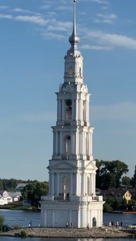 The video shows a tall, white, multi-tiered bell tower on a small island in a body of water. The tower has a spire topped with a gold cross. The lower portion of the tower is wider and features arches and columns. There are people visible on the island at the base of the tower and on a lower platform. Some people are standing and looking at the tower, while others appear to be walking. Small boats are visible in the water. In the background, there is a shoreline with buildings and trees. The sky is blue with some scattered clouds. The video is presented as a series of still frames, suggesting it may be a time-lapse or a sequence of photographs.