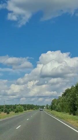 The video shows a car driving on a paved road surrounded by lush greenery under a bright blue sky dotted with fluffy white clouds. The road is marked with white lines and has a single lane in each direction. A white car is visible in the distance, driving in the same direction. On the left side of the road, there are power poles and trees, while the right side is lined with dense green trees. A triangular warning sign with a red border and a black symbol inside is visible on the right side of the road. The sun appears to be high in the sky, suggesting it is daytime, likely late morning or early afternoon. The weather appears to be clear and pleasant. The overall scene depicts a scenic drive through a rural or semi-rural landscape in Novotroitskoye, Russia. There is no discernible text in the video, aside from a faint, partially visible sign on the left side of the road which appears to be a road sign.