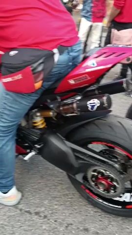 The video shows a red Ducati motorcycle at what appears to be a gathering or event. A man wearing a red shirt and jeans is straddling the bike. The motorcycle is customized with a distinctive exhaust system. Several people are standing around, some taking photos or videos with their phones. The bike has red accents on the wheels and various stickers. The environment looks like an outdoor parking lot or street, with other bikes and pedestrians visible in the background. The engine makes a loud sputtering sound when it is turned on.