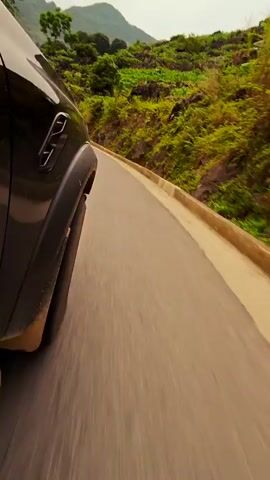 A car navigates a winding, paved mountain road. The camera is positioned low, near the rear tire of the vehicle, providing a view of the road ahead and the surrounding landscape. Green, tree-covered mountains rise in the distance under a slightly overcast sky. The road is lined with concrete barriers on the passenger side, separating the road from the rocky, vegetation-covered slope. A sign that reads "Beautiful View" is visible on the side of the road.
