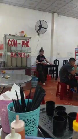 The video captures a scene inside a Vietnamese restaurant, likely a "Com-Pho" (rice and Pho) establishment in Hanoi, judging by the sign on the glass-fronted cabinet displaying rows of bowls. A young woman wearing a black t-shirt and patterned pants walks towards the camera, carrying a bowl in her hands, possibly delivering food to a customer. The restaurant setting includes simple furniture, such as plastic stools and tables. A man is seated at one of the tables, eating. In the foreground, there's a table with various condiment containers including those for chili sauce. Also seen are containers holding chopsticks and straws. A black fan is mounted on the wall behind the bowl display case. The general environment seems simple and functional.