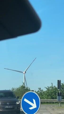 The video shows a view from inside a car, likely from the driver's perspective. The main subjects are a wind turbine in the distance and a blue traffic sign with a white arrow pointing to the right. The wind turbine has three blades and is turning slowly. The sky is clear and blue. In the foreground, the car's sun visor is partially visible at the top of the frame. Other cars are seen in the background moving towards the right. The video appears to be taken during the day.
