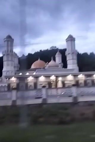 The video begins with a medium shot of a mosque with a large dome and two tall minarets. The mosque is white and has decorative arches. The sky is cloudy and dark, suggesting it is either dawn or dusk. A line of trees and a hill are visible behind the mosque. As the video progresses, the camera appears to be moving, possibly from inside a vehicle, indicated by the slight blur and the way the mosque recedes into the distance. Power lines are visible in the upper portion of the frame. A dark-colored car is seen driving past the mosque on a road. The video ends with a close-up shot of the entrance area of the mosque, showing more of its intricate architectural details. The overall scene is serene and hints at a religious or spiritual atmosphere.