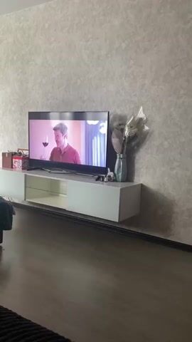 The video shows a living room setting with a large flat-screen TV mounted on the wall. On the TV, a man is drinking from a wine glass. The TV is sitting on a long white TV stand with a glass shelf. To the right of the TV, there is a vase with white flowers. The wall is a light gray with a textured pattern. The floor is made of dark wood planks. The room appears tidy and well-lit. The video is stable, and the focus is on the TV and surrounding area.