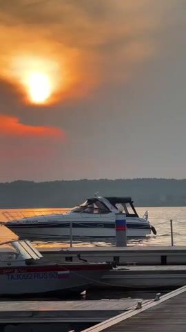 The video shows a sunset over a body of water, likely a lake or river. The sun is bright and low in the sky, casting a warm orange glow through the clouds. Several boats are docked in the foreground, with one larger white yacht prominently featured. The water is calm, reflecting the colors of the sunset. The scene appears to be peaceful and serene, likely a marina or a recreational area.