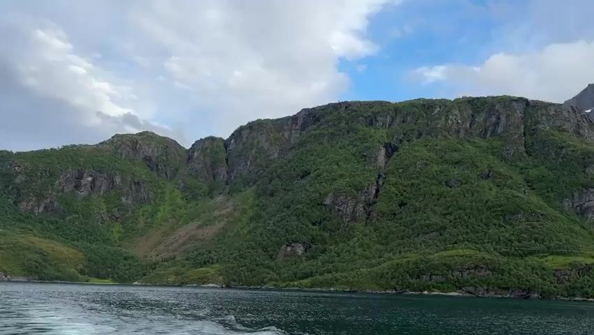 The video shows a wide shot of a body of water, likely a fjord, with steep, green, tree-covered mountains on the right side. The water is dark and has a visible wake behind the camera, indicating movement. In the distance, a long, concrete bridge spans across the water, connecting to a small, rocky island and continuing towards the mainland. The bridge has a distinctive curved design. Further back, snow-capped mountains rise majestically, their peaks partially obscured by clouds. The sky is overcast with patches of blue peeking through. The overall scene is serene and majestic, showcasing a natural landscape with human infrastructure.