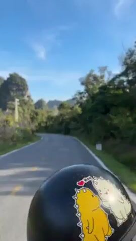 The video, shot from a first-person perspective, shows someone riding a motorcycle on a paved road. The rider is wearing a black helmet decorated with yellow cartoon figures and red hearts. The road curves gently, with trees and vegetation lining both sides. In the distance, there is a prominent rock formation, possibly a mountain or cliff, covered with trees. A white utility pole is visible on the left side of the road. The sky is clear and blue.  A car passes by in the opposite direction in the background.