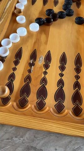 A hand rolls two dice onto a wooden backgammon board. The dice show a 2 and a 1. The board is made of light-colored wood with dark brown designs. White and black game pieces are arranged on the board. The board is on a stone surface.