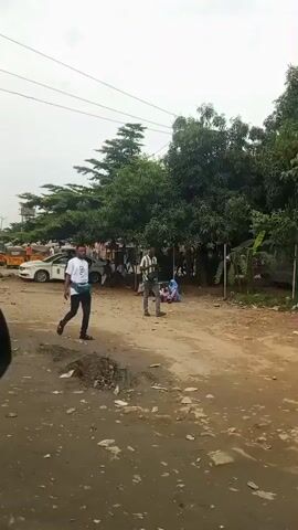 The video shows a dirt road with several people and vehicles. In the foreground, a person in a white t-shirt and a fanny pack walks across the road. To the left, there is a white car with its doors open and a person standing next to it. Further down the road, there are several other cars, including what appear to be taxis or buses. There are also trees and foliage on either side of the road, and power lines overhead. In the background, several people are sitting under a makeshift shelter. The overall scene appears to be a busy roadside area in a developing country.