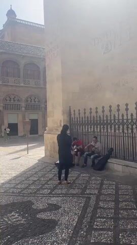 The video pans across a public square, likely in a European city, given the architecture. Several people are visible. In the foreground, a person in a black coat stands facing away from the camera, appearing to observe or interact with two men sitting on the ground playing guitars. One guitarist wears a red shirt, and the other wears a gray shirt. Both have guitar cases beside them. The ground of the square is paved with pebbles arranged in a mosaic pattern. Behind the musicians, a wrought-iron fence separates the square from a building with a large, ornate facade. Further in the background, more people walk through a cobblestone street lined with buildings featuring balconies and arched windows. The architecture suggests a historic European setting, with warm-toned stone buildings and intricate decorative elements. Sunlight casts shadows, indicating it is daytime. The overall atmosphere seems relaxed, with street musicians adding to the ambiance.