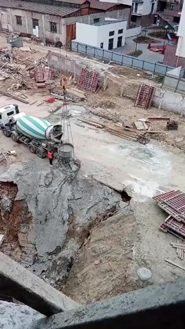 A construction site is shown from an elevated perspective. A large concrete mixer truck is parked next to a large hole in the ground. A crane is lifting a concrete bucket from the truck, presumably to be lowered into the hole. A construction worker in an orange vest is standing near the truck and the edge of the hole. The site is cluttered with various construction materials and debris. In the background are buildings and a partially visible playground. The day appears overcast.