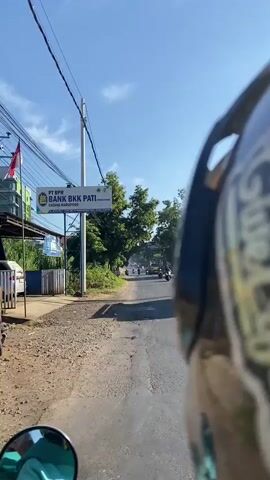 The video shows a view from the perspective of someone riding a motorcycle on a paved road. The camera is looking forward and slightly to the right, showing the rider's helmet in the foreground. On the left side of the road, there is a sign that reads "PT BPIS BANK BKK PATI (PERSERO)". A utility pole with many wires is also visible on the left. Beyond the sign and pole, there are trees and vegetation. The road ahead is mostly empty except for a few distant vehicles. The sky is blue with some clouds, and the sun is shining, casting shadows on the road. The overall scene appears to be a rural or semi-urban area during the daytime.