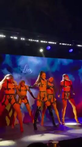 Okay, here is a concise description of the video:

The video shows a group of five dancers performing on a stage. They are all wearing skimpy outfits with harnesses. The lighting is primarily blue and red, with spotlights illuminating the dancers. Behind the dancers, there is a large screen displaying an image of fiery red clouds. The dancers are moving in sync, performing energetic choreography. The video appears to be shot from the audience, and some audience members can be seen in the foreground. There is also music playing in the background.