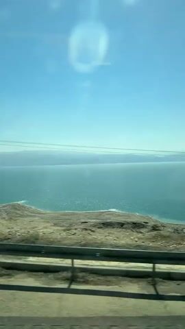A vehicle is moving along a road with a guardrail, next to a land area that drops down to a body of water. The water is turquoise, and the land is brown. The sky is clear and blue with some light reflections in the car window. Power lines are visible on the horizon above the water.