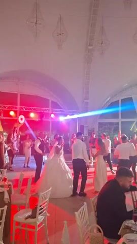 The video shows a wedding reception in a large, white, tent-like venue with arched windows. In the center of the frame, a bride in a white wedding gown and a groom in a dark suit are dancing. They are surrounded by other wedding guests, some of whom are also dancing. A band or DJ setup is visible on a stage in the background with bright red and blue lights. Tables with white chairs are set up in the foreground, some with food and drinks on them. The lighting is dim and atmospheric, with spotlights illuminating the dance floor. The overall mood appears festive and celebratory.