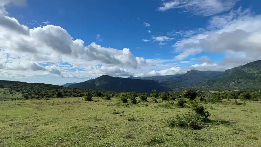 The video shows a scenic view of a grassy field with mountains in the background under a partly cloudy sky. The field has low shrubs scattered throughout. A dirt path appears to the right of the frame. The mountains are covered in greenery, with some shrouded in clouds. The sky is blue with patches of white clouds. The wind is slightly audible.