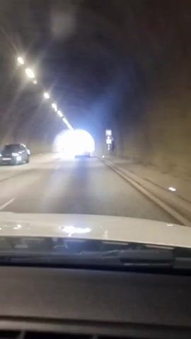 The video shows a point-of-view shot from inside a vehicle traveling through a tunnel. The tunnel is illuminated by overhead lights and a bright light source at the end, suggesting the exit. The tunnel walls are made of concrete and appear to be curved. There are traffic signs visible on the right side of the tunnel. A few other vehicles are present, including one in the distance ahead and another in the opposing lane. The camera view is focused on the road ahead, with the dashboard of the vehicle visible in the foreground. The overall impression is of transit through an underground passage.
