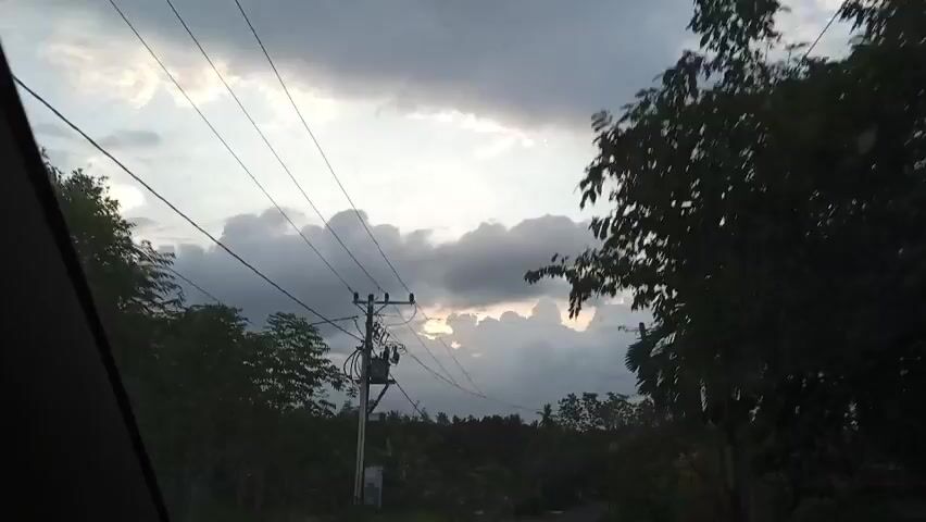 The video shows a view of a cloudy sky through the window of a moving vehicle. Power lines and poles run across the view of the sky. The view through the window also shows trees and foliage. The overall lighting suggests it's either early morning or late afternoon.