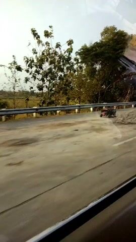 The video captures a view from a vehicle in motion. The foreground is dominated by a paved road with some discoloration and debris. Beyond the road, a metal guardrail separates the road from a grassy embankment lined with trees. The trees vary in size and foliage density. In the background, a field is visible, and the sky is clear with a slight haze. A small red object, possibly a piece of machinery or equipment, is partially visible on the embankment. The overall scene suggests an outdoor environment, likely a rural or semi-rural roadside.