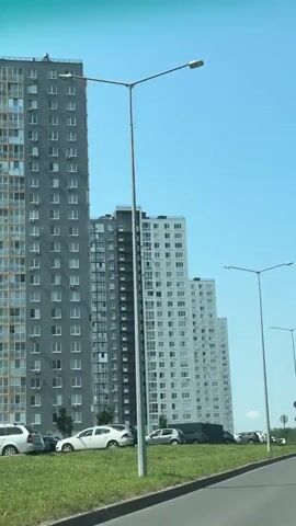 The video shows a view from a moving vehicle looking towards a row of tall apartment buildings. The buildings are modern and appear to be mostly light gray with some sections featuring yellow or darker gray elements. They are multi-story structures with numerous windows and balconies. In front of the buildings, there is a grassy area with a row of parked cars. A paved road curves in the foreground, and streetlights are visible along the side of the road. The sky is clear and blue. The footage appears to be captured during the daytime, likely on a sunny day. There are no people visible in the video. The camera perspective suggests the video was taken while traveling parallel to the buildings.