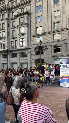 A group of people are gathered in an outdoor plaza in front of a large, ornate building. A few people are standing on a raised platform or stage, some of whom are holding a Venezuelan flag. One person on the stage appears to be speaking into a microphone. Another person on the stage is playing a guitar. A banner with text and logos is visible on the stage. The crowd of onlookers are facing the stage, some are holding cell phones up, presumably to record. The people in the foreground are facing away from the camera, towards the stage. The building behind the stage has multiple stories with windows and architectural details. There is a lamppost visible in the center of the plaza. The ground is paved with reddish-brown stones.