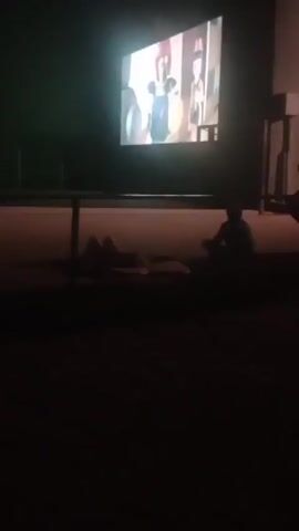 A dimly lit outdoor space at night. A large projection screen displays an animated movie. In the foreground, two people are lying down on the ground, and a third person is sitting cross-legged. The movie appears to be a children's animated film. The overall impression is of a casual outdoor movie viewing experience.