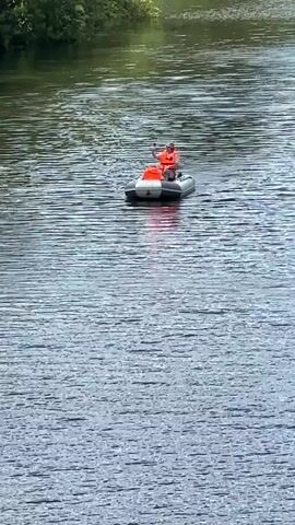 A person wearing an orange life vest is in an inflatable raft on a body of water. The person is holding a paddle and appears to be rowing. The raft is gray and white. The water is calm with ripples. Trees line the distant bank of the water.