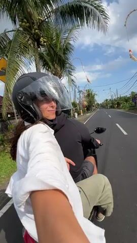 A man and woman are riding on a scooter down a paved road. The woman is in front, wearing a black helmet and a white shirt. The man is behind her, wearing a black hoodie and light-colored pants. They are both looking forward, and the woman appears to be smiling. The road is lined with palm trees and other vegetation. There are buildings visible on either side of the road, and some decorative banners are hanging from poles along the roadside. The sky is blue with some white clouds. The video appears to be a first-person perspective, possibly recorded by the woman.