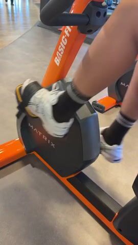A close-up shot focuses on a person's legs and feet as they pedal an orange and black stationary exercise bike. The bike has the brand name MATRIX visible on its console. The person is wearing white athletic shoes with black and yellow accents, black socks, and athletic shorts. The pedals are secured with straps. The person's legs are in motion, indicating they are actively exercising. The background is slightly blurred, showing a gym floor and other gym equipment, including an orange frame of another piece of equipment and part of a person in the far left background.