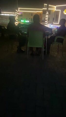 Three people are sitting at a small table outdoors at night. The people are silhouetted, so their details are not visible, but it appears they are men. They are sitting in plastic chairs that are green and white. The table is small and metal. They appear to be talking. Behind them is a building with yellow neon lights and some trees. The ground is paved.
