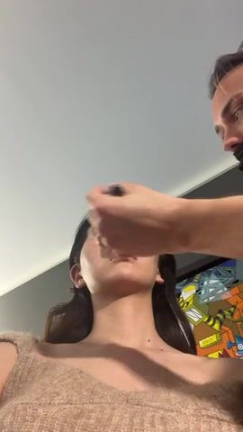 A low-angle shot shows a young Asian woman with her face tilted up, receiving makeup. A man with light skin and a black face mask is applying makeup to her face using a brush. The woman has dark hair, some of which is pulled back into a bun, and she's wearing a fuzzy tan sweater. Behind her is a colorful, cartoon-style mural. The camera slowly zooms in on the woman's face, revealing glossy lips and subtle makeup. Background chatter is audible.