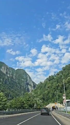 The video shows a view from inside a vehicle traveling on a highway. The highway is paved and has two lanes visible. There are guardrails along the side of the highway. Mountains covered in lush green trees are visible on the left and right sides of the road. The sky is a bright blue with scattered white clouds. A tunnel entrance is visible on the right side of the road, with cars driving towards it. A blue truck with white writing on its side passes the vehicle from behind.