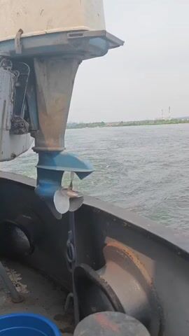 This video shows the propeller of a boat in the water. The propeller is made of metal and is turning, creating a wake in the water. The boat is moving through the water, and there is a shoreline in the background with some buildings and industrial structures. The sky is cloudy. The video is shot from the perspective of someone on the boat, looking over the side.