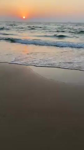 The video depicts a tranquil sunset scene at a beach. The sun, a bright orange sphere, hangs low on the horizon, casting a warm glow across the sky. Gentle waves roll onto the sandy shore, their foamy edges creating a contrasting white line against the darker sand. The beach is empty, with no people visible in the frame. The water reflects the sunset's colors, enhancing the overall serene and peaceful atmosphere.