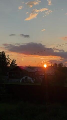 This media file captures a tranquil sunset scene in Emel'yanovo, Russia. The sun, a brilliant orb, is positioned just above the horizon, casting a warm, golden glow across the sky. It's surrounded by a palette of soft oranges, pinks, and purples, blending seamlessly into the calming blue of the twilight sky. Wispy clouds drift lazily, catching the last rays of sunlight and adding depth to the celestial canvas.

The foreground is dominated by the silhouette of a dark wooden fence, behind which lush green foliage thrives. Beyond the fence, a cluster of houses with dark roofs and some with greenhouses are visible, suggesting a rural or suburban setting. A tall wooden utility pole stands prominently to the right of the frame, its wires stretching across the sky and contributing to the overall atmosphere of a quiet, lived-in landscape. Trees, dark and leafy, frame the scene on the left and in the background.

The time of day is clearly sunset, evidenced by the low position of the sun and the vibrant colors in the sky. The weather appears clear and calm, ideal for enjoying the sunset. The overall mood conveyed is one of peace, serenity, and the gentle winding down of a day in the Russian countryside. There is no visible text in the media file.
