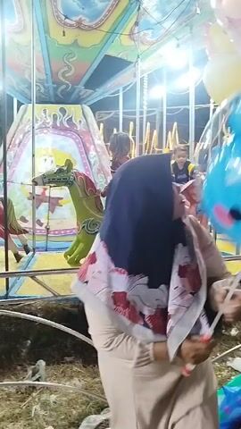The video shows a carousel with children riding on various animal figures. In the foreground, a woman wearing a hijab and a patterned dress is partially visible from the side. In the background, a young girl in a red jacket and jeans is riding a green horse. Next to her, a child is riding a pink elephant. Further back, another child is seen on a giraffe. The carousel is brightly lit with colorful decorations on the canopy and the central structure, which features a depiction of SpongeBob SquarePants. The ground beneath the carousel appears to be covered in dry grass or straw. The overall scene depicts a nighttime amusement park setting.
