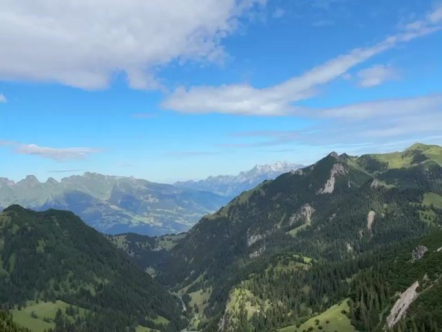 A panoramic drone shot captures a vast mountain range under a bright blue sky dotted with white clouds. The camera pans slowly across the landscape, revealing layers of green, forested mountains fading into distant, hazy peaks. The foreground features steep, rocky slopes covered with sparse green vegetation and dark evergreen trees. A winding dirt path is visible on the right side of the frame. The scene is devoid of any people or man-made objects, emphasizing the natural, rugged beauty of the alpine environment. The overall impression is one of serene, expansive wilderness.