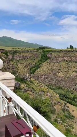 The video shows a panoramic view of a rocky canyon with green vegetation. The canyon walls are steep and layered, with evidence of rock formations and erosion. Lush green trees and bushes grow along the canyon floor and on the slopes. In the distance, there are rolling hills and a cloudy sky. The camera is positioned on a balcony with a white railing, suggesting an elevated viewpoint overlooking the natural landscape. There are no people visible in the video. A small herd of animals, possibly cows, can be seen grazing on the lower slopes of the canyon. The overall impression is one of a vast and rugged natural environment.