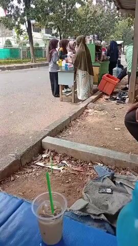A street-side scene with several people near a small stand selling drinks. In the foreground, a plastic cup with a green straw contains a frothy brown beverage, possibly iced coffee. The cup rests on a blue surface, likely a table or seating. Behind it is a patch of dry dirt with scattered debris including a gray fabric scrap. Beyond the dirt is a concrete curb and an asphalt road. On the right side of the road, a woman wearing a dark hijab and a patterned skirt is reaching into a red container near the stand. Another woman in a lighter hijab stands behind a small table with various items on it, including bottles and what appears to be fruit. Several other individuals are visible further down the street, some near the stand and others walking away. Trees and buildings are visible in the background. The overall impression is a casual, outdoor marketplace setting.
