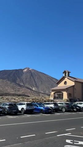 The video shows a parking lot filled with various cars. In the background, a large mountain is visible under a clear blue sky. To the right of the mountain, there is a building that resembles a church or a visitor center. The camera pans slowly from left to right, revealing more cars and the landscape. The parking lot is paved and marked with white lines.