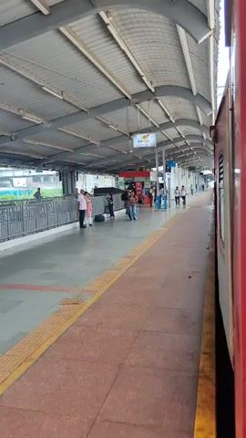 The video shows a train at a train station platform. The train is red and white and is on the right side of the frame. The platform is gray with a yellow tactile paving strip along the edge. There are people waiting on the platform, some with luggage. A roof covers the platform. A screen is visible hanging from the roof.