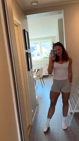The video features a young woman taking mirror selfies inside what appears to be a brightly lit hallway or entryway of a home, possibly located in Bergen, Norway.

In the initial shot (0:00-0:02), the woman stands before a large mirror, capturing her reflection with a light-colored smartphone held in her right hand. She has medium brown hair, worn down, with sunglasses pushed up onto her head. She is dressed in a white form-fitting tank top, light grey athletic shorts, and white crew socks. A light pink scrunchie is visible on her left wrist. She offers a gentle smile to the camera. The background, reflected in the mirror, shows a light-colored wooden floor, a white wall adorned with two vertically hung black-framed pictures, and a partially visible white, collapsible drying rack to her right. Through an open doorway further back, a sunny room with a large window is discernible, revealing green trees and residential buildings outside, along with a white chair.

After a brief, blurry transition (0:03), the video cuts to a second selfie shot (0:04-0:06). In this closer and higher-angle view, the woman is seen extending her arm to take another mirror selfie. She displays a broad, cheerful smile, revealing dimples, and her left hand is casually placed on her stomach. She wears the same attire and accessories. The background reflections confirm the identical indoor setting, with the window, outdoor scenery, and drying rack still present.

The overall atmosphere is bright and casual, with the woman appearing happy and comfortable in her home environment. The interior is modern and tidy, and the outdoor view suggests a residential area. No visible text is present in the video.