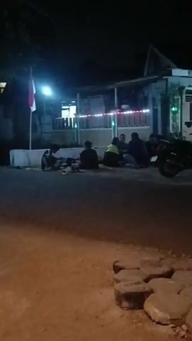 The video shows a group of people sitting on the ground in front of a building at night. There is a flagpole with a flag that appears to be red and white. The building has lights on it, and there are motorcycles parked nearby. The people are gathered in a circle and appear to be socializing. The ground is paved, and there are some decorative stones in the foreground. The overall atmosphere is casual and relaxed.