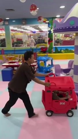 A man is pushing a red toy car with a child inside. The man is wearing a brown t-shirt and black pants. The child is wearing a grey outfit. They are in a playground with a carousel, slides, and other toys. The floor is covered with colorful foam mats. The walls are decorated with colorful banners and posters. There are shelves with toys in the background. The lighting is bright and artificial.