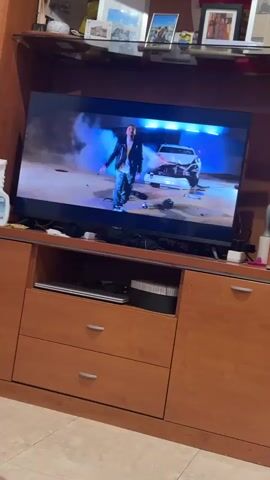 The video displays a television screen showing snippets of a music video. The first scene features a man in a leather jacket standing next to a crashed car in a smoky, blue-lit environment. The second scene shows a man and a woman sitting at a table with a patterned background. The third scene returns to the man standing by the crashed car, gesturing with his hands. The final scene shows a man and a woman looking at each other by a body of water. The television is placed on a wooden entertainment center with drawers.