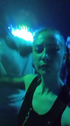 The video shows a person filming themselves at an outdoor concert with a stage and lights in the background. The person is close to the camera and the background is blurry. The concert appears to be at night, with blue and green lighting dominating the scene. There are other people in the crowd around the person.