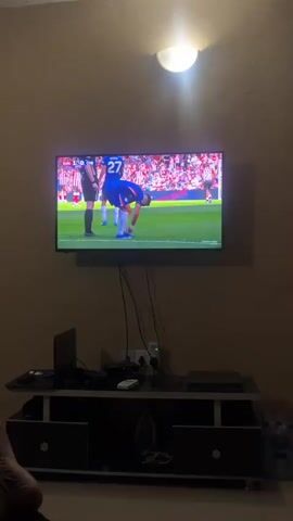 Here's a description of the video:

The video shows a room with a TV mounted on the wall, playing a soccer game. The lighting in the room is dim, with a single ceiling light illuminating the scene. Below the TV, there is a black and white TV stand. A laptop, a game console, and various electronic accessories are on the stand. 

The TV screen displays a live soccer match with players on a green field. A player in a blue uniform is bent over, seemingly adjusting his sock or shin guard. Another player in a blue uniform, wearing the number 20 is standing straight. Several players wearing striped red and white jerseys surround them. A densely packed stadium crowd is visible in the background. The score on the TV indicates "SUN 1-0 CHE".

In the lower left corner of the frame, there is a glimpse of someone's foot, suggesting the camera is held by a person sitting or lounging in the room.